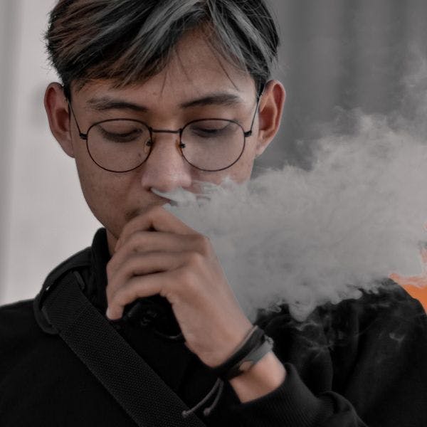 A person with glasses and a black shirt with a white smoke coming out of his mouth.
